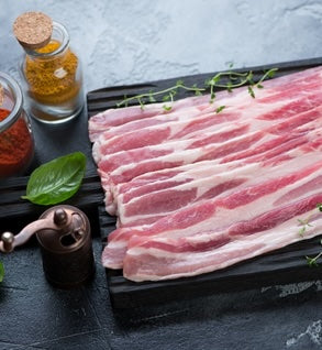Raw pork bacon strips laid out on a dark surface, with visible seasonings and a pepper mill.