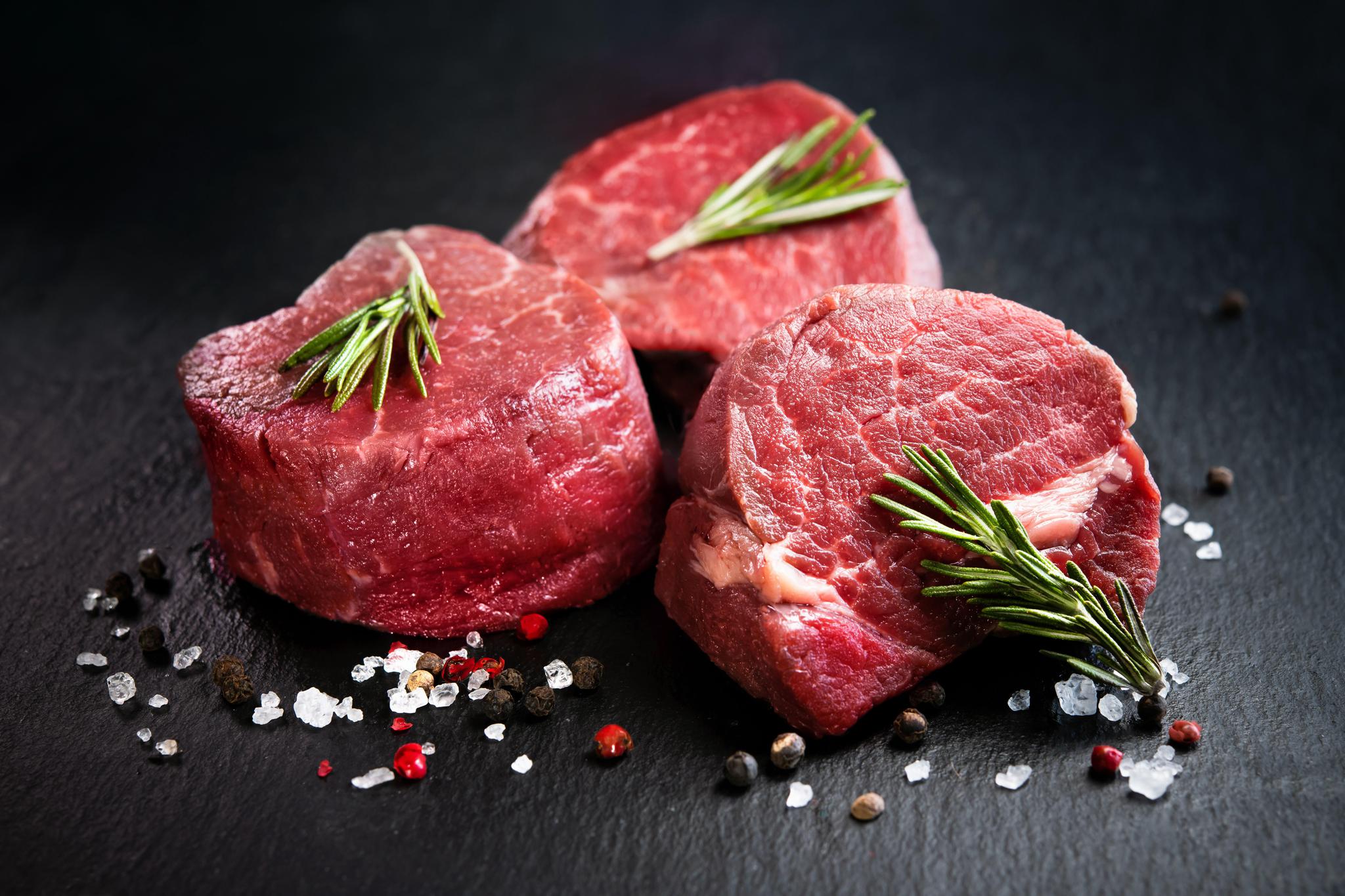 Three filet mignon steaks on a dark surface with salt, pepper, and rosemary leaves scattered around them.