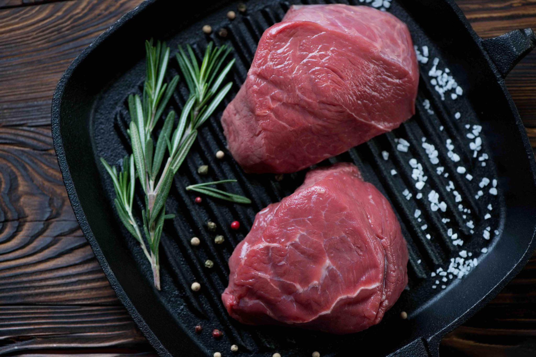 Two filet mignon steaks on a dark surface with salt, pepper, and rosemary leaves scattered around them.